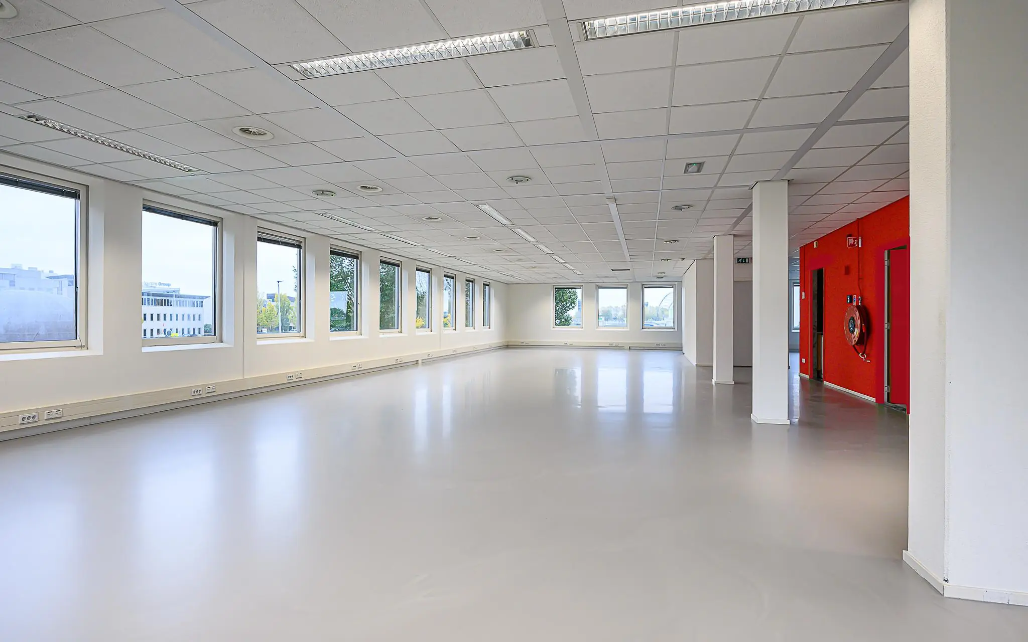 Lichte, lege kantoorruimte aan de Rivium Westlaan met grote ramen en een rode accentmuur.